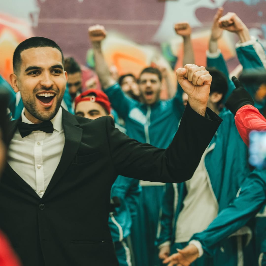 DHM en smoking lève le poing avec enthousiasme au milieu des participants en tenue verte lors de sa vidéo Squid Game IRL